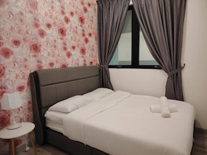 Een net opgemaakt bed met witte lakens en een opgerolde handdoek, een klein nachtkastje met een lamp, en een raam met gordijnen.