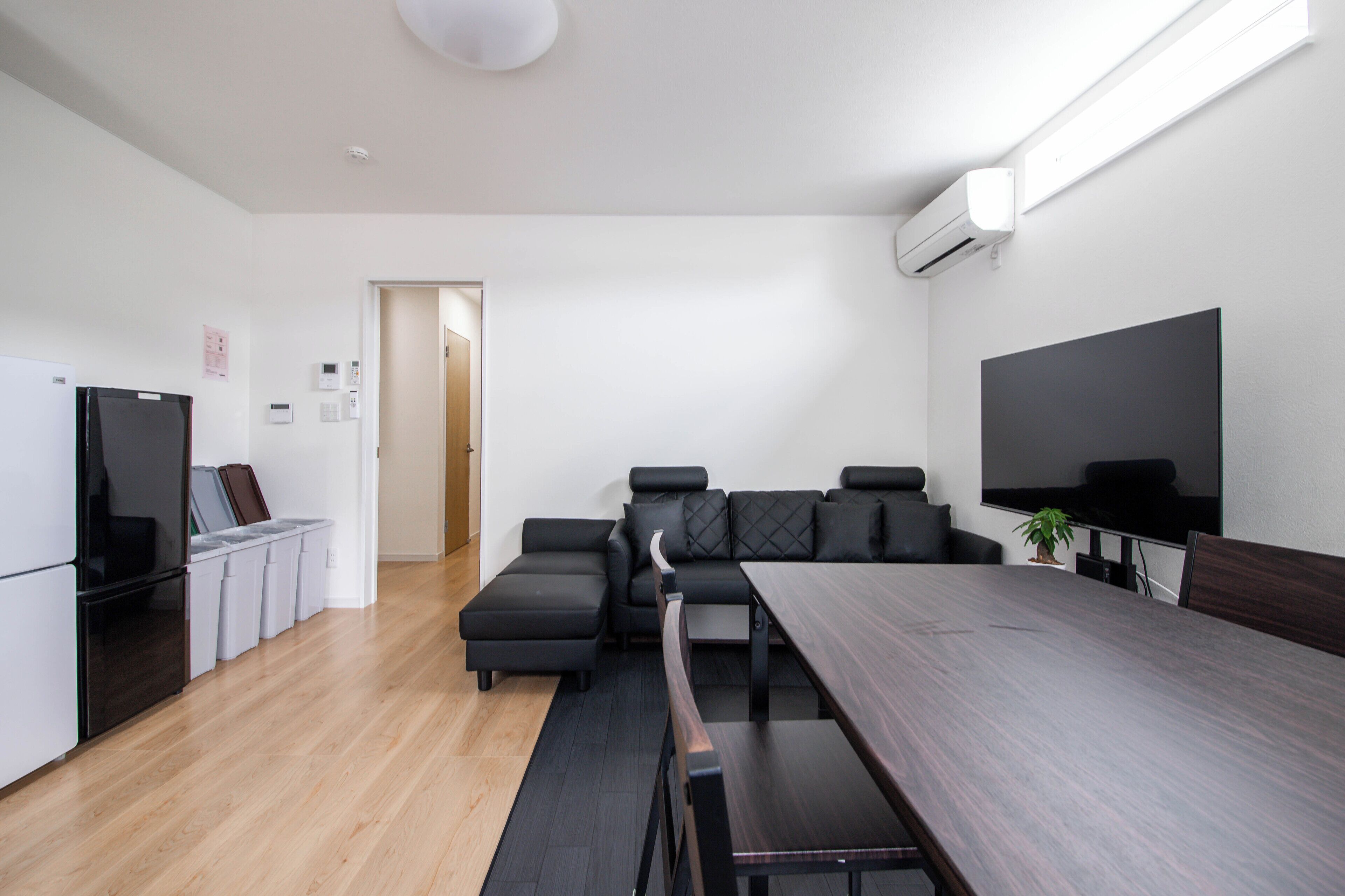 A modern kitchen with a refrigerator, stove, and trash bins. There is a dining area with a long table and benches. A television is mounted on the wall.