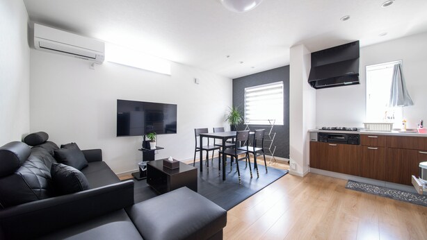 Una sala de estar moderna con un sofá negro, un comedor con mesa y sillas, una cocina con armarios de madera y un televisor de pantalla plana montado en la pared.
