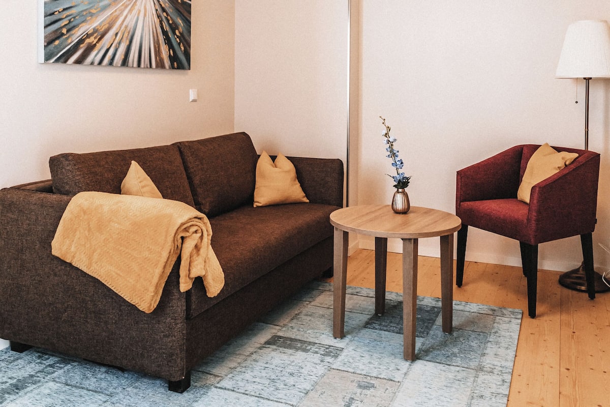 A modern living room with a brown sofa, a red armchair, a small round table with a vase, and a large abstract painting on the wall.