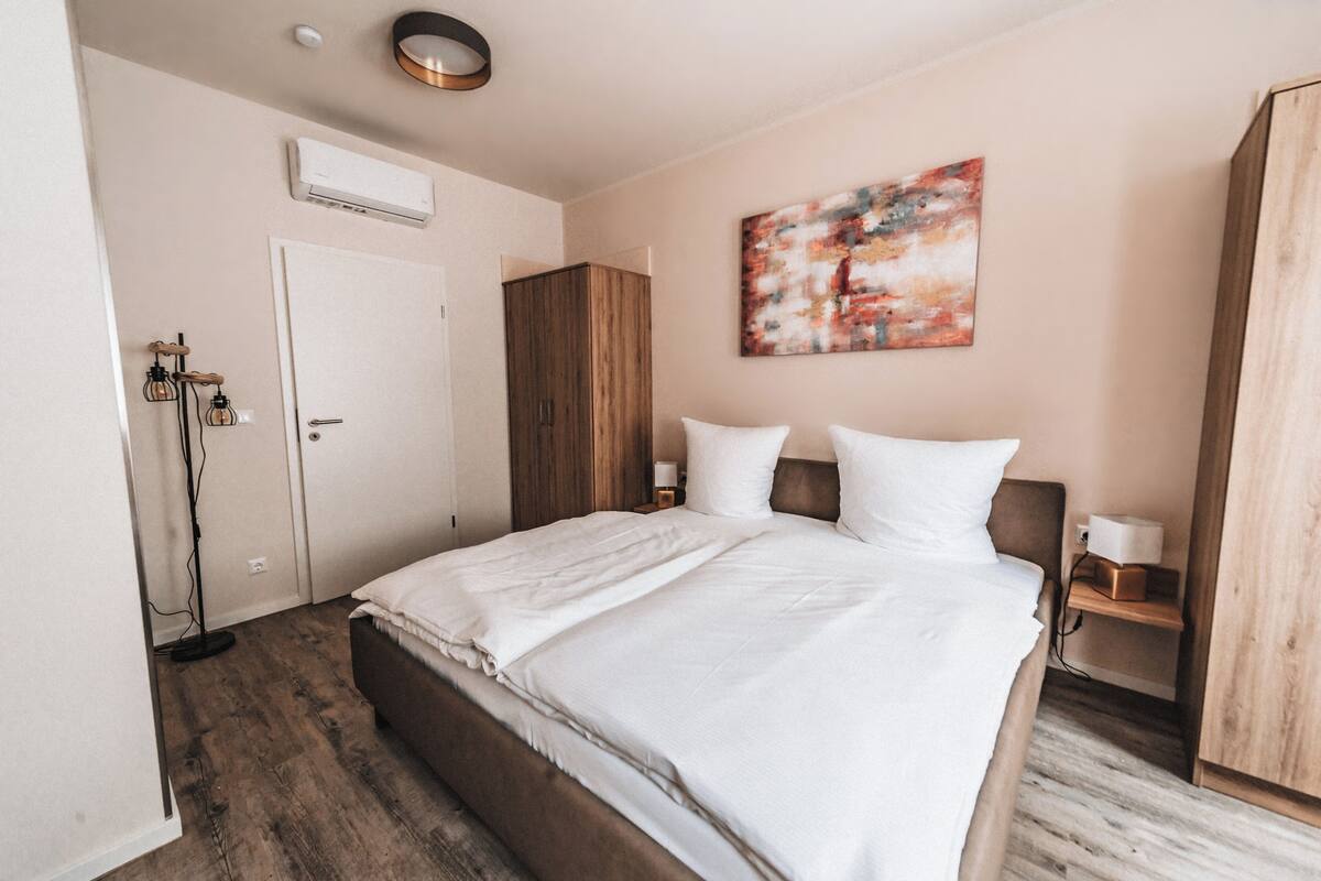 A bedroom with a large bed, a wooden wardrobe, a painting on the wall, and a white door.