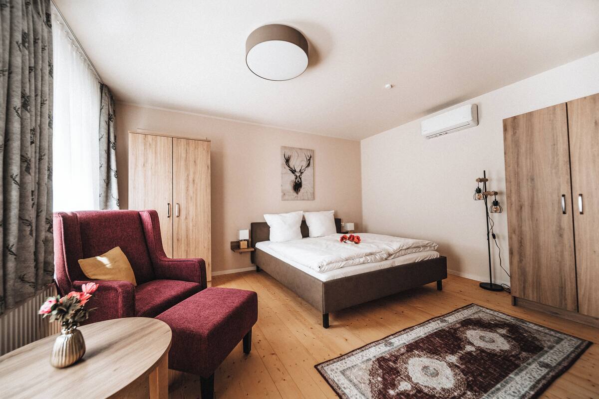 A modern bedroom with a bed, a red armchair, a wooden wardrobe, and a small table with a vase.