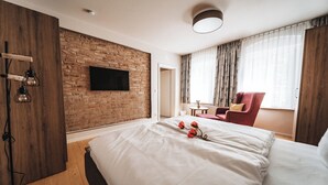 A hotel room with a brick wall, a flat-screen TV, a bed with white linens, a red armchair, and a small table with a vase. - Fontane Apartments (Schorfheide)