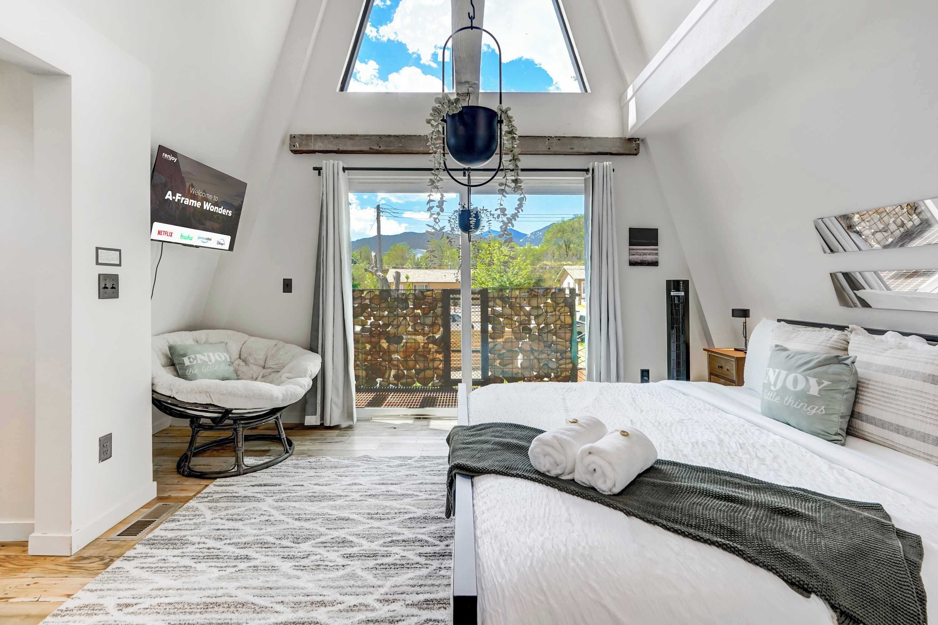 Un dormitorio con cama, una silla, una planta colgante y vistas al exterior.