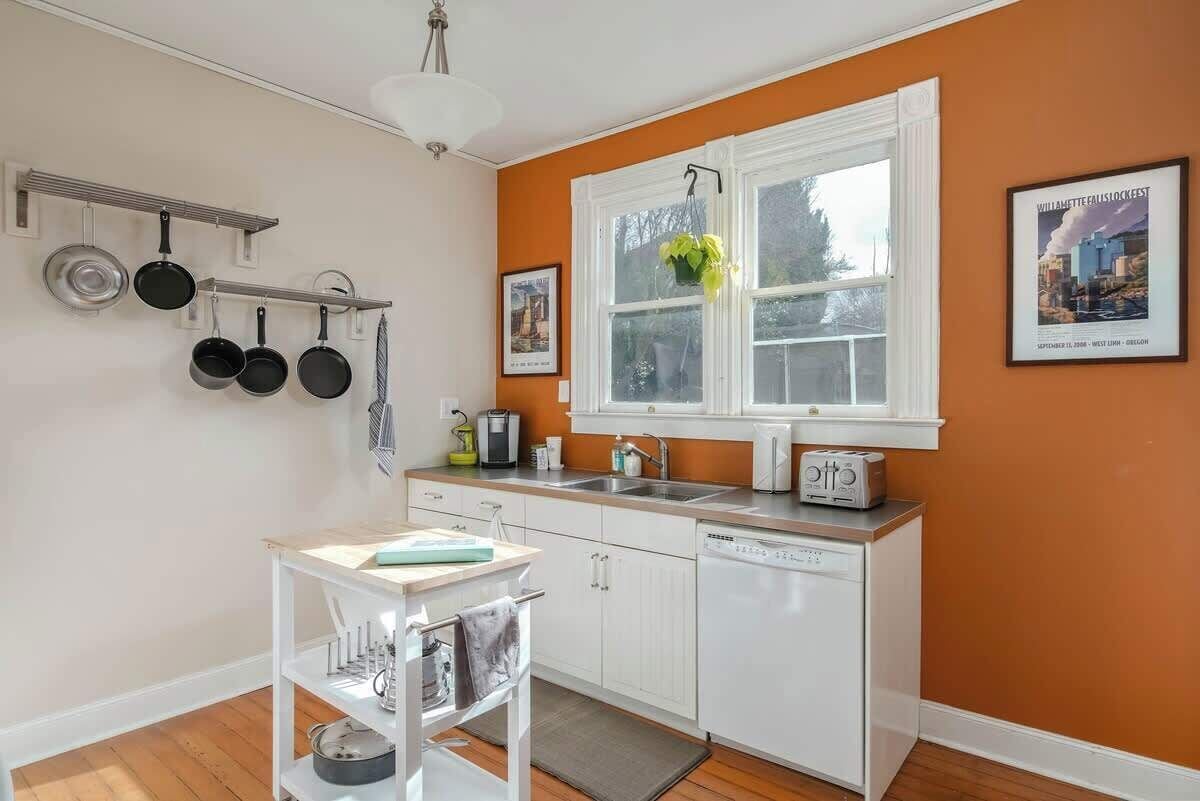 Une cuisine avec des meubles blancs, un plan de travail blanc, un porte-ustensiles suspendu, une fenêtre avec une plante et un grille-pain.