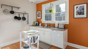 Une cuisine avec des meubles blancs, un plan de travail blanc, un porte-ustensiles suspendu, une fenêtre avec une plante et un grille-pain.