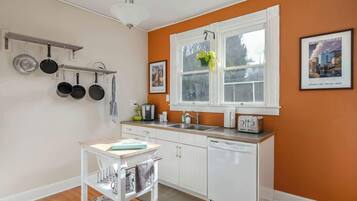 Une cuisine avec des meubles blancs, un plan de travail blanc, un porte-ustensiles suspendu, une fenêtre avec une plante et un grille-pain.