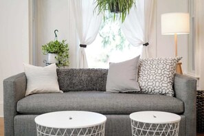 Ein graues Sofa mit gemusterten Kissen, zwei runde weiße Tische mit geometrischen Mustern, eine hängende Pflanze und eine Lampe.