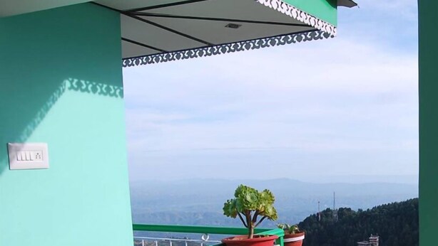A balcony with a laptop, a green mug, and a notebook on a wooden table. There are potted plants on a railing and a view of a mountainous landscape. - Jagatram Niwas (Dharamshala)
