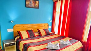 A bedroom with a wooden headboard, a bed with patterned bedding, a nightstand, and a red and white striped curtain. - Jagatram Niwas (Dharamshala)
