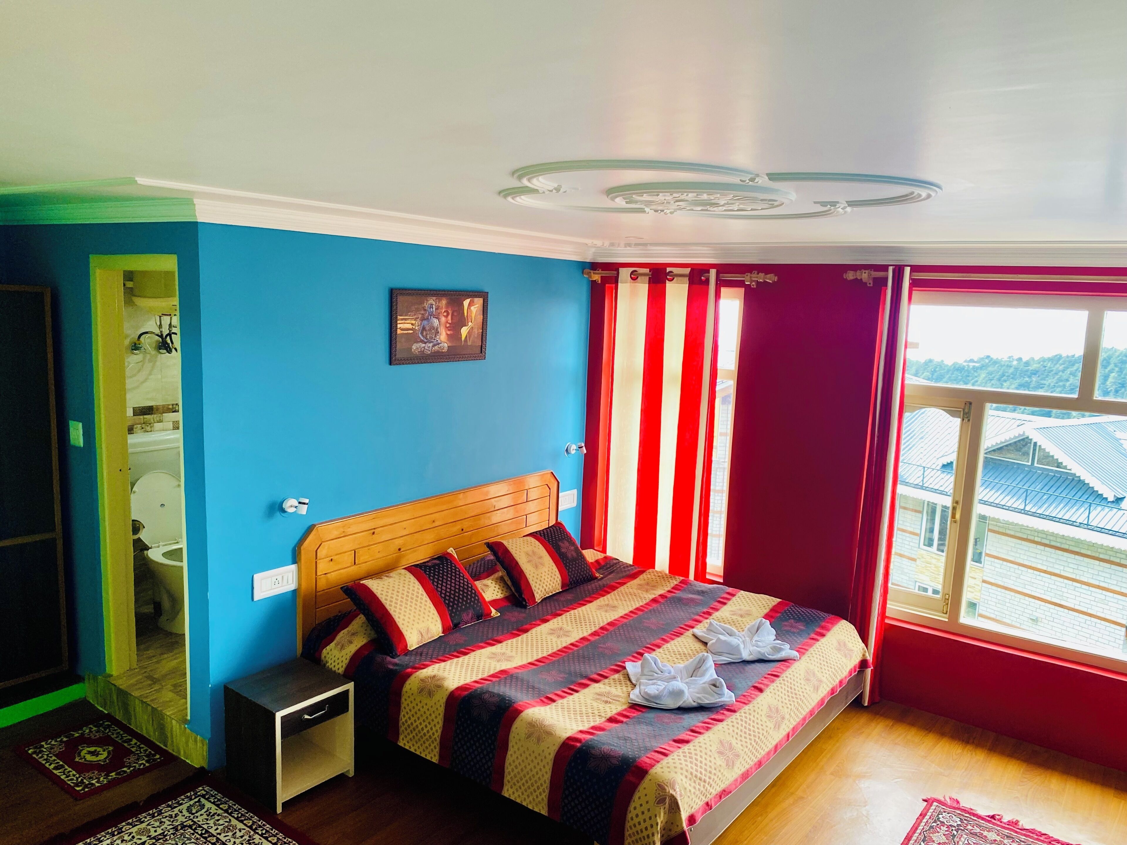 A bedroom with a bed, a window with curtains, a bathroom visible through an open door, and a wall-mounted picture.