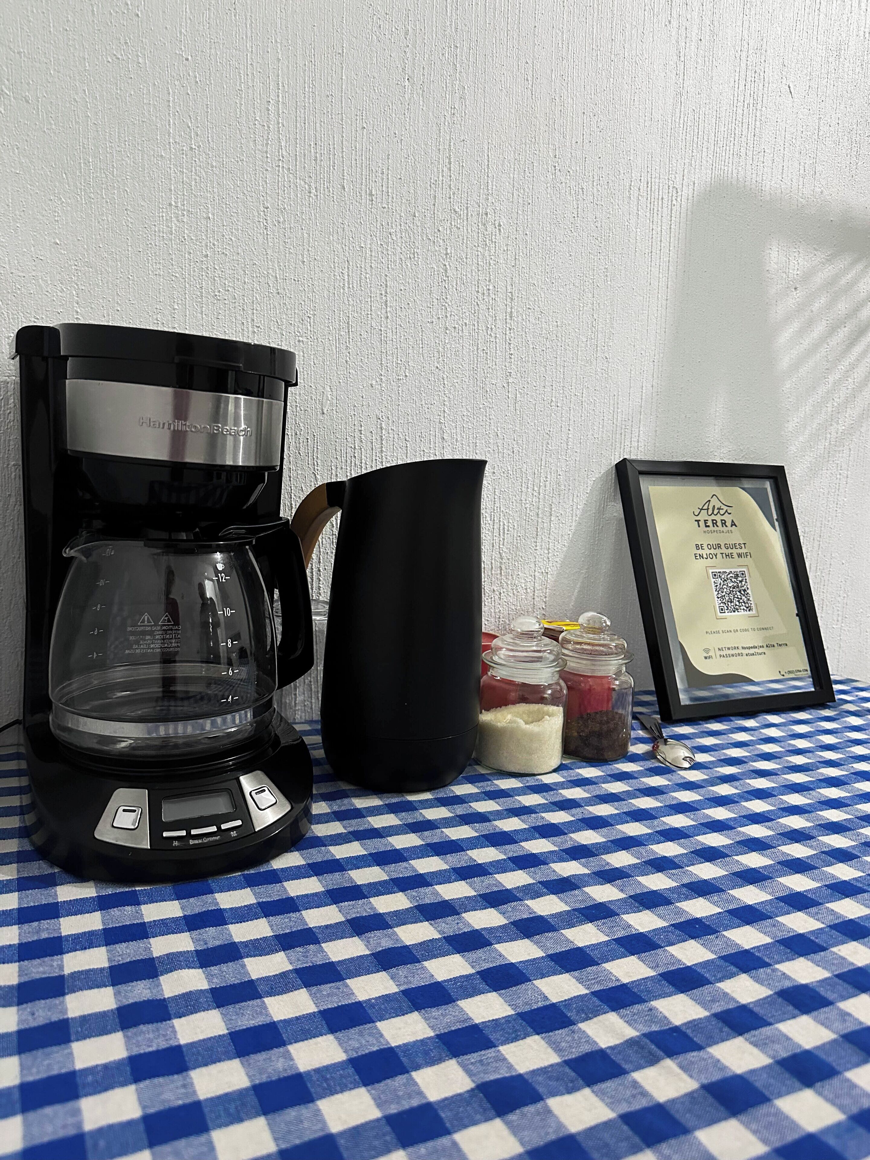 Een koffiezetapparaat, een zwarte karaf, potten suiker en kruiden, en een bord met een QR-code op een geruit tafelkleed.