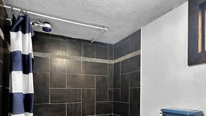 A bathroom with dark tiled walls, a shower with a striped curtain, a blue toilet, and a small rug on the floor. - Hospedajes Alta Terra (Quetzaltenango)