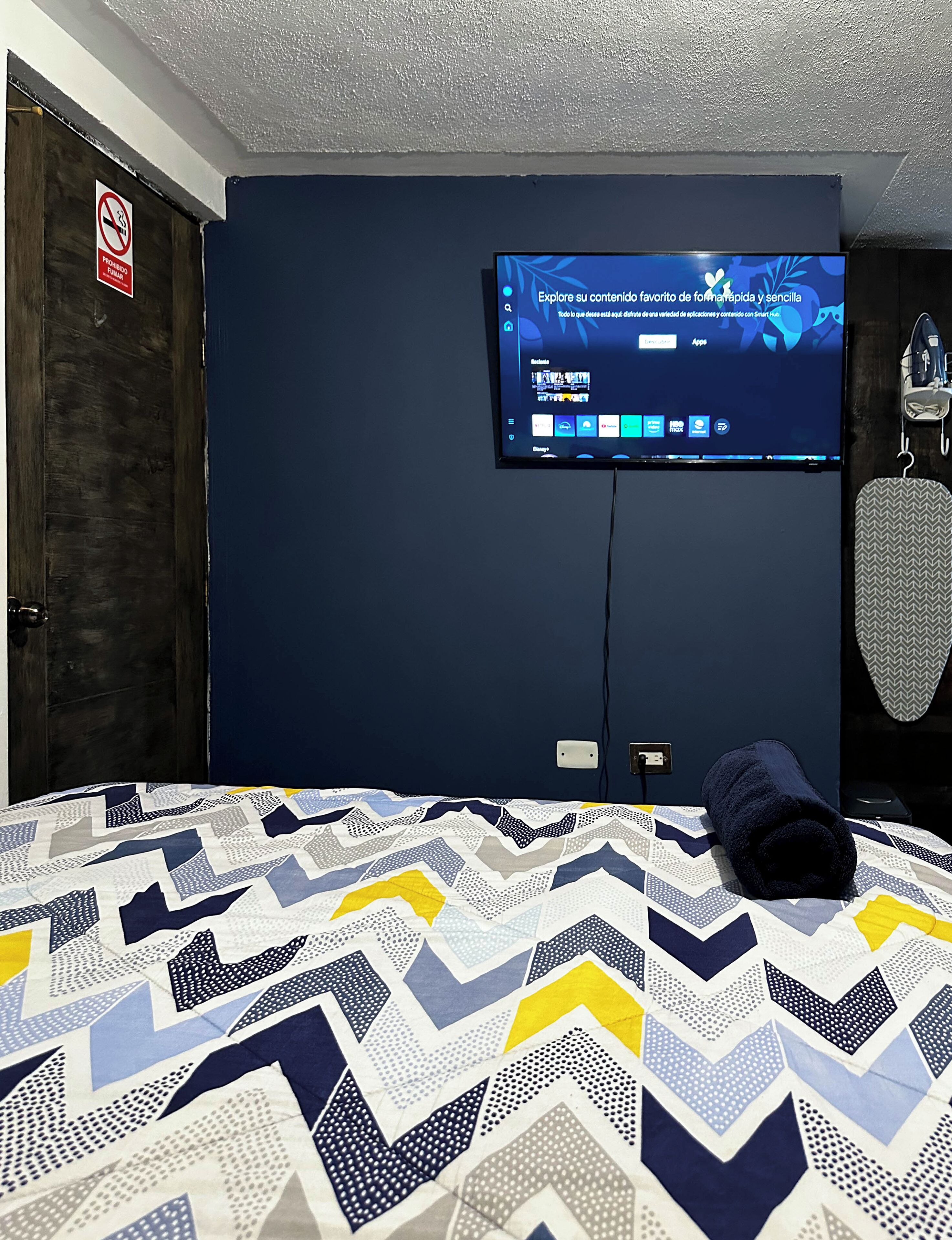 A hotel room with a bed, a television mounted on the wall, and a door with a no smoking sign.