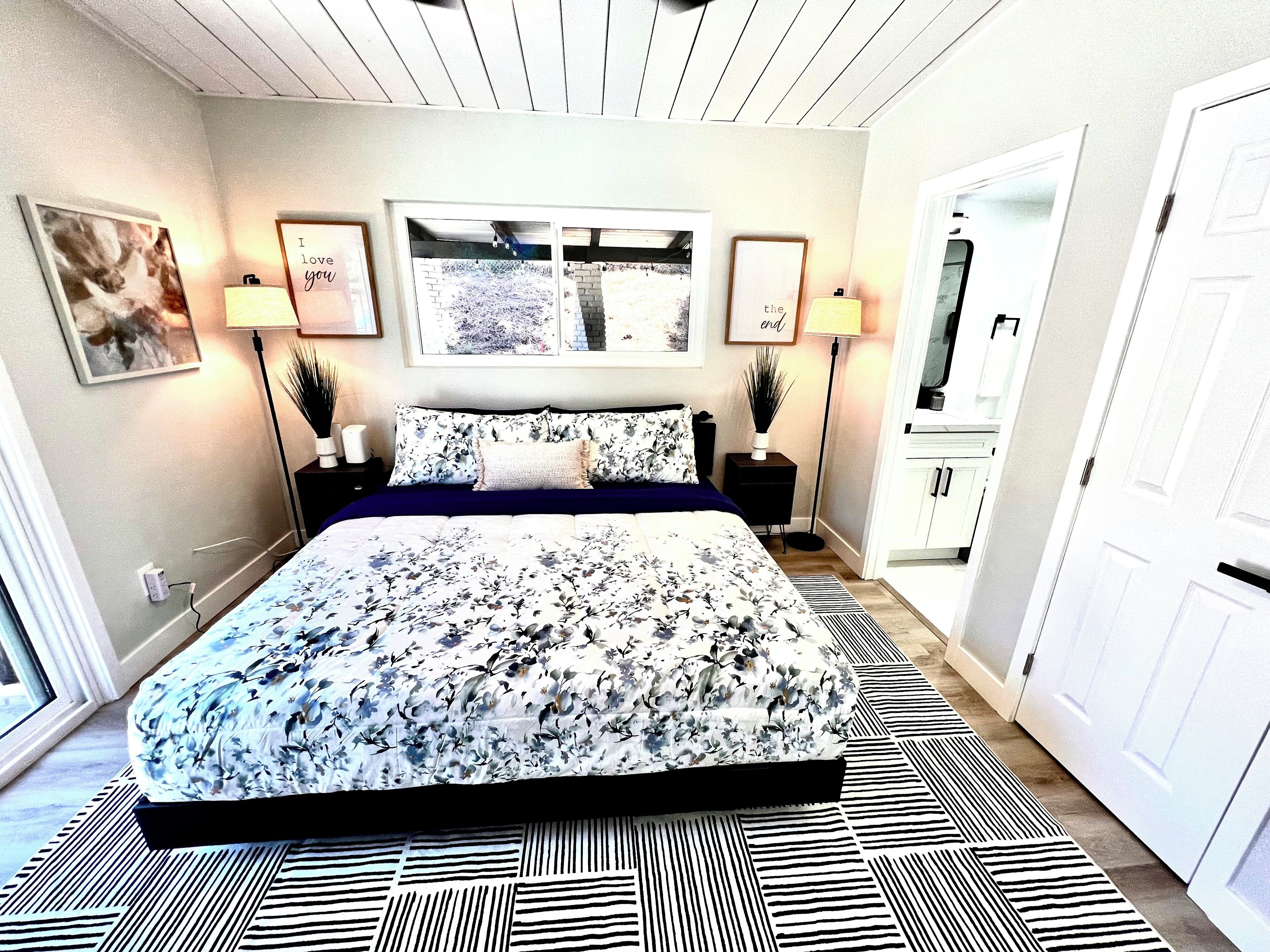 Une chambre à coucher avec une couvre-lit à motifs floraux, deux lampes de chevet, une citation encadrée accrochée au mur et une vue sur le couloir.