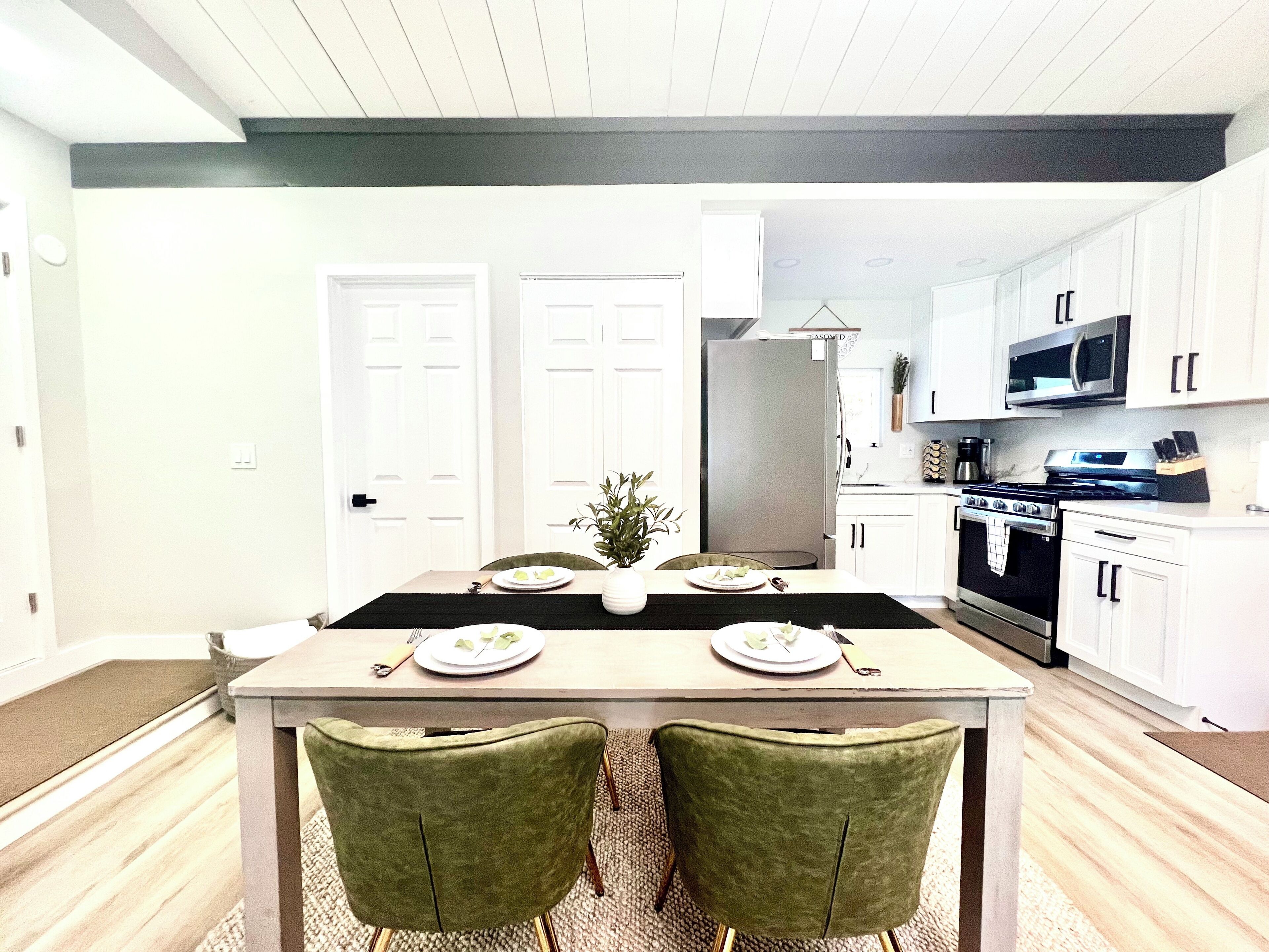 Une cuisine moderne avec des meubles blancs, un réfrigérateur en acier inoxydable et un four à micro-ondes. On y trouve une table à manger avec des chaises et une petite plante en pot.