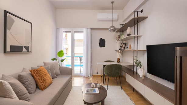 A modern living room with a sofa, coffee table, and TV. There is a dining area with a chair and table. A plant is visible, and the room has a large window. - Lycabettus Modern Retreat (Athina)