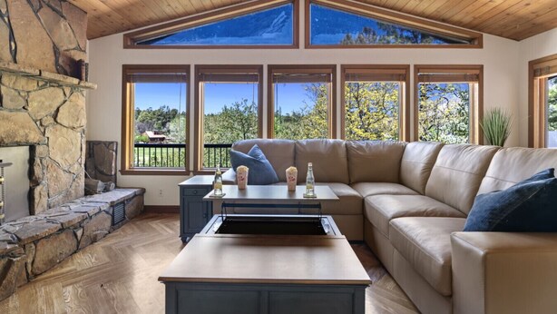 A spacious living room with a stone fireplace, a large window with a view of trees, a beige sectional sofa, and a wooden ceiling. - Xbox*Ping Pong* Pop-a-shot*Game Room* (Flagstaff)