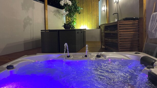 Ein Whirlpool mit Wasserstrahlen und blauer Beleuchtung, umgeben von Holzmöbeln und einem Blumentopf.