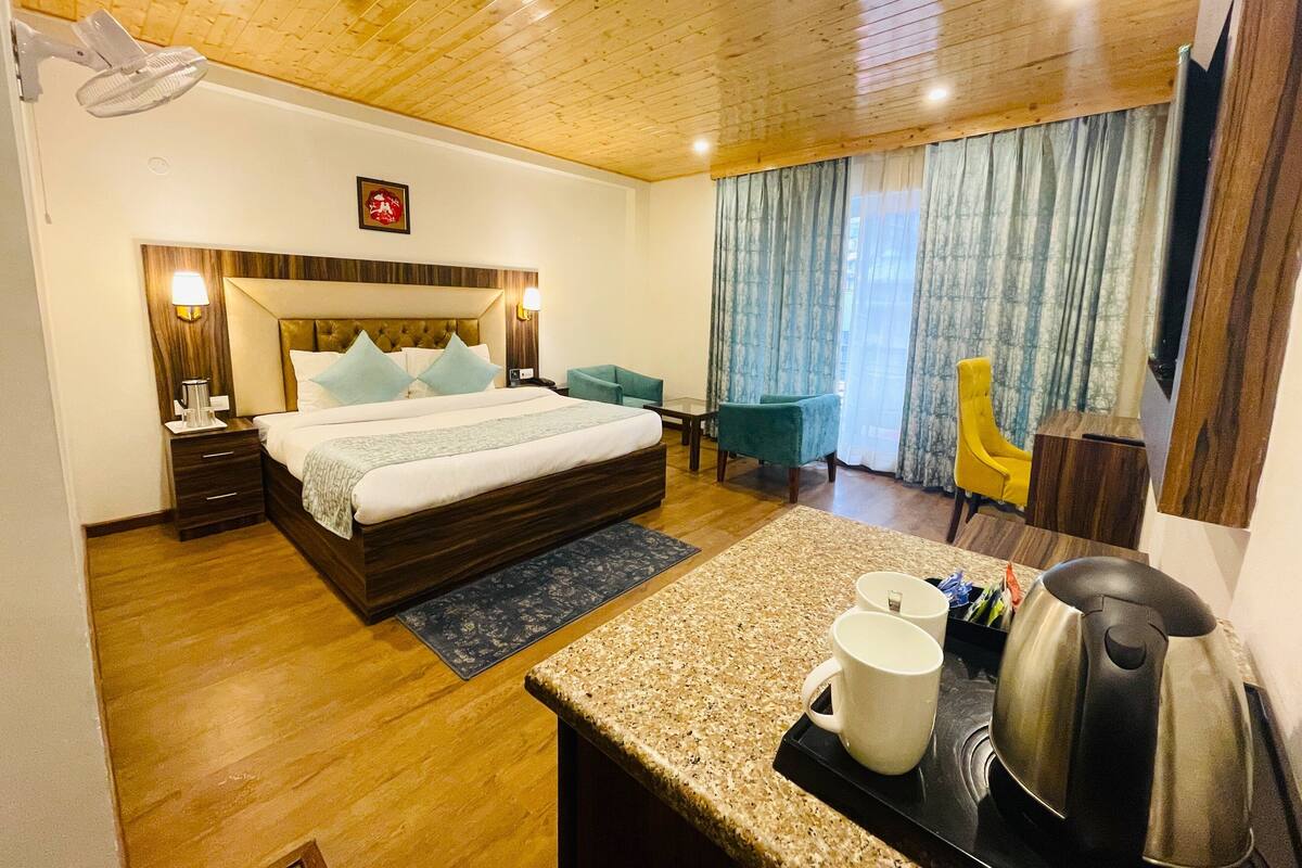 A hotel room with a wooden ceiling, a bed with a headboard, a desk with a kettle, and a chair.