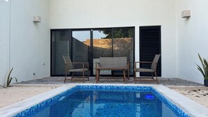 Un área de piscina exterior moderna con piscina con azulejos, dos sillas de mimbre y una pequeña mesa.