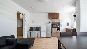 A modern kitchen with white appliances, wooden cabinets, and a dining area with a dark table and black sofa. - Apollo (Hakodate)