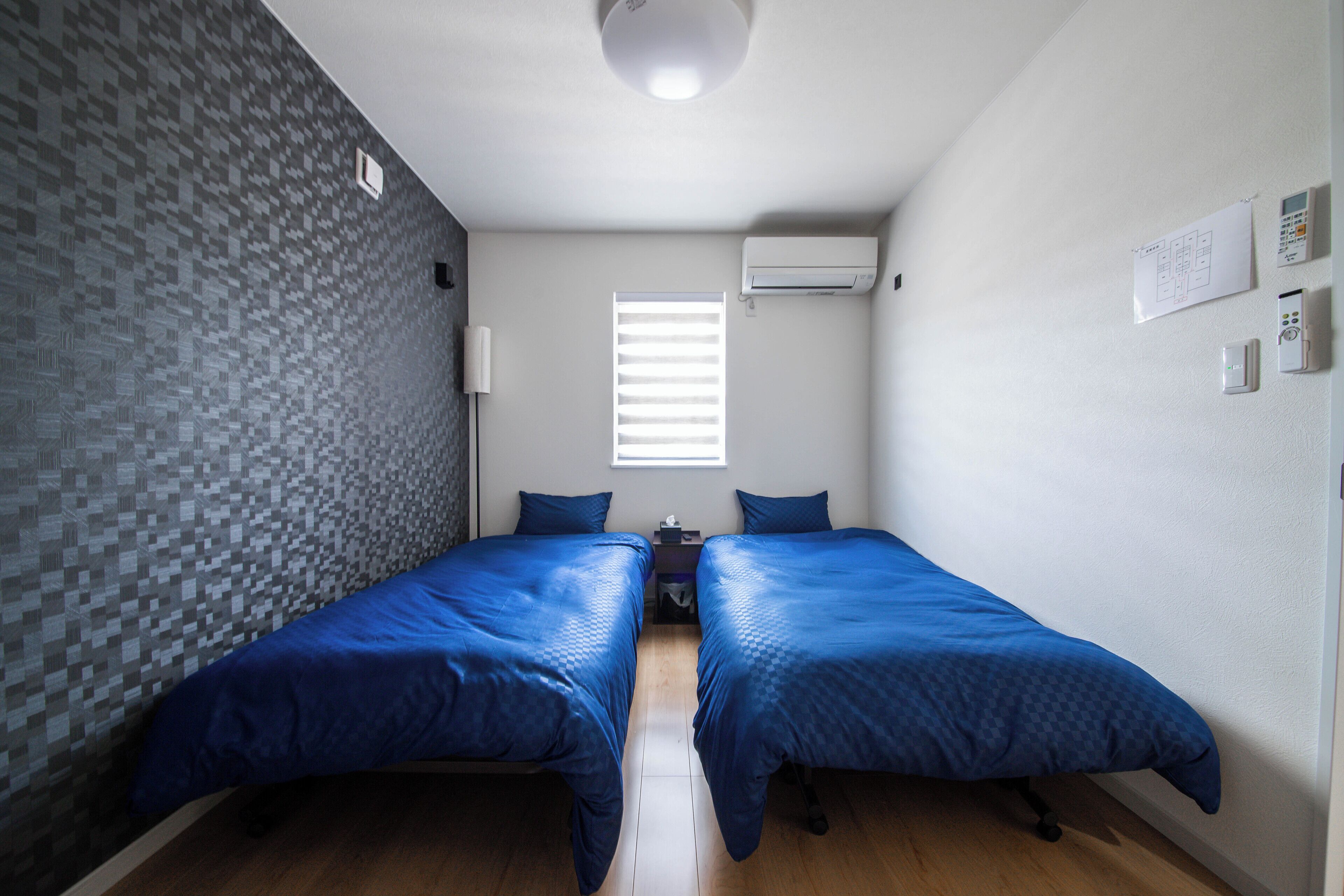 A small, modern room with two single beds, a window with blinds, an air conditioning unit, and a wall-mounted control panel.