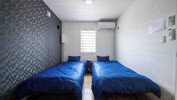 A small, modern room with two single beds, a window with blinds, an air conditioning unit, and a wall-mounted control panel.