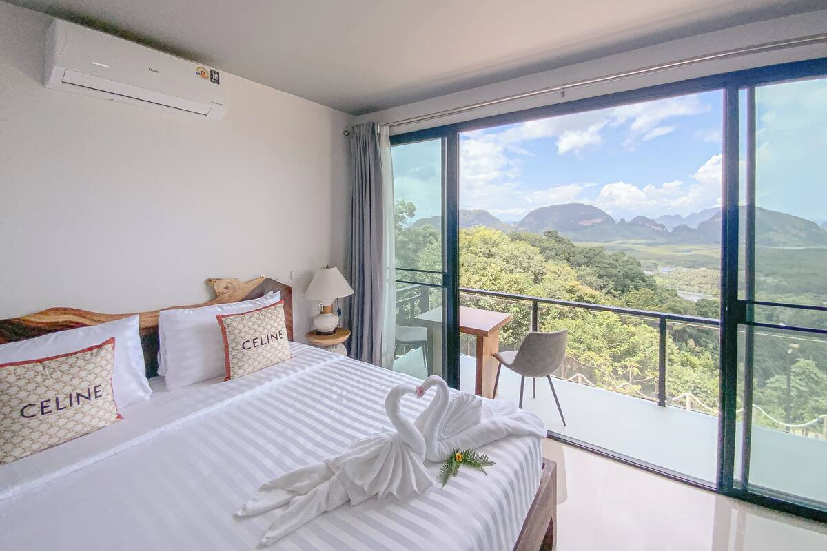 A hotel room with a large bed, a view of mountains, a balcony with a table and chair, and a ceiling fan.