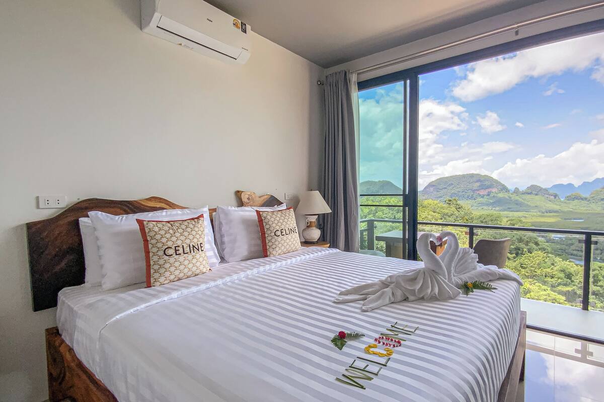 A hotel room with a large bed, a view of a mountainous landscape, and a balcony with a table and chairs.