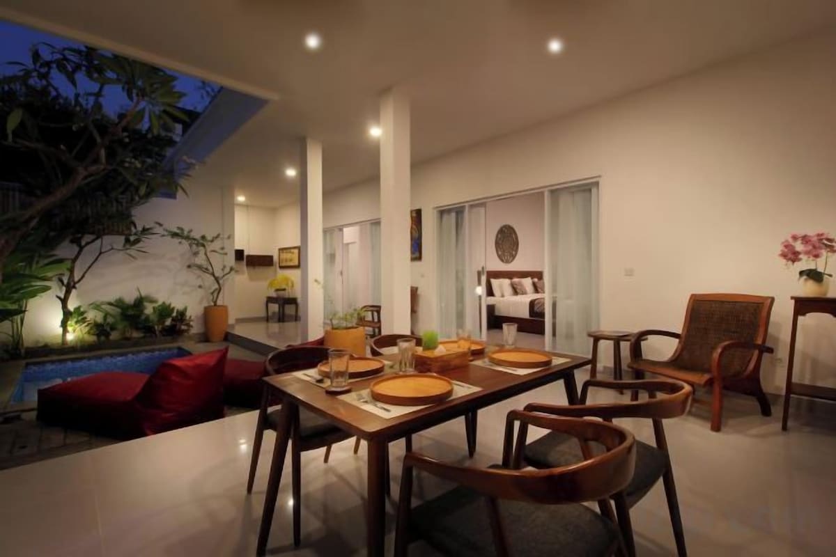 A modern living room with a dining area, a pool, and a bedroom visible through an open door.