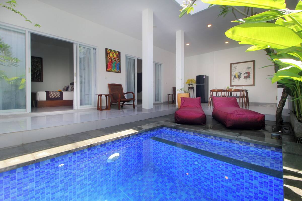 Area piscina interna moderna con vasca blu a mosaico, porte scorrevoli che conducono a un soggiorno e un tavolo da pranzo con sedie.
