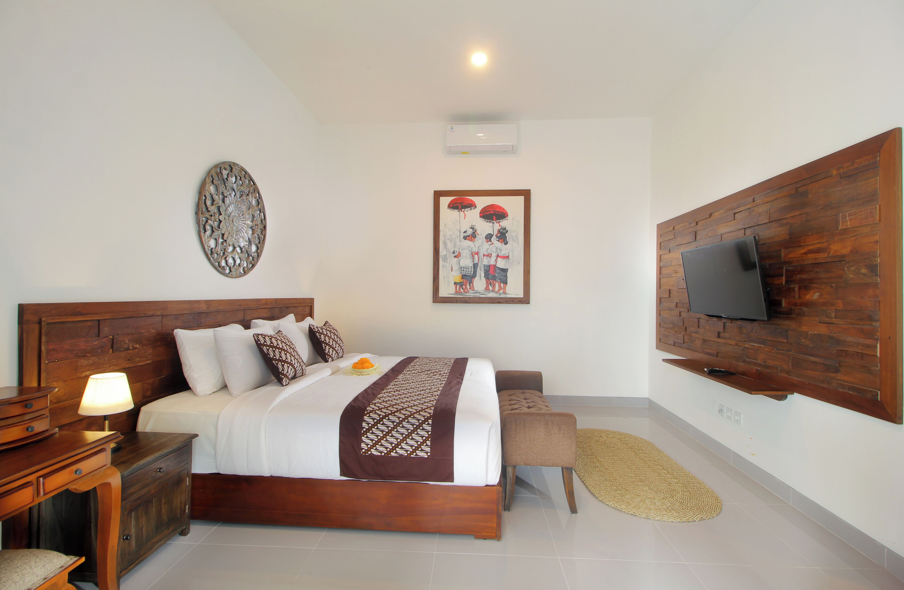 A bedroom with a wooden headboard, a bed with white bedding and patterned pillows, a bedside table with a lamp, a wall-mounted TV, and a decorative wall piece.