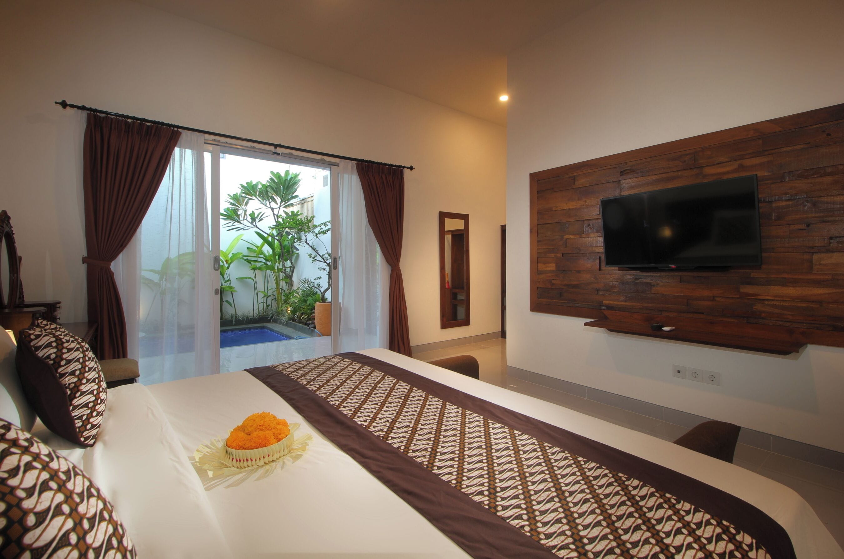 A hotel room with a bed, a TV mounted on a wooden wall, and a view of a pool and greenery outside.