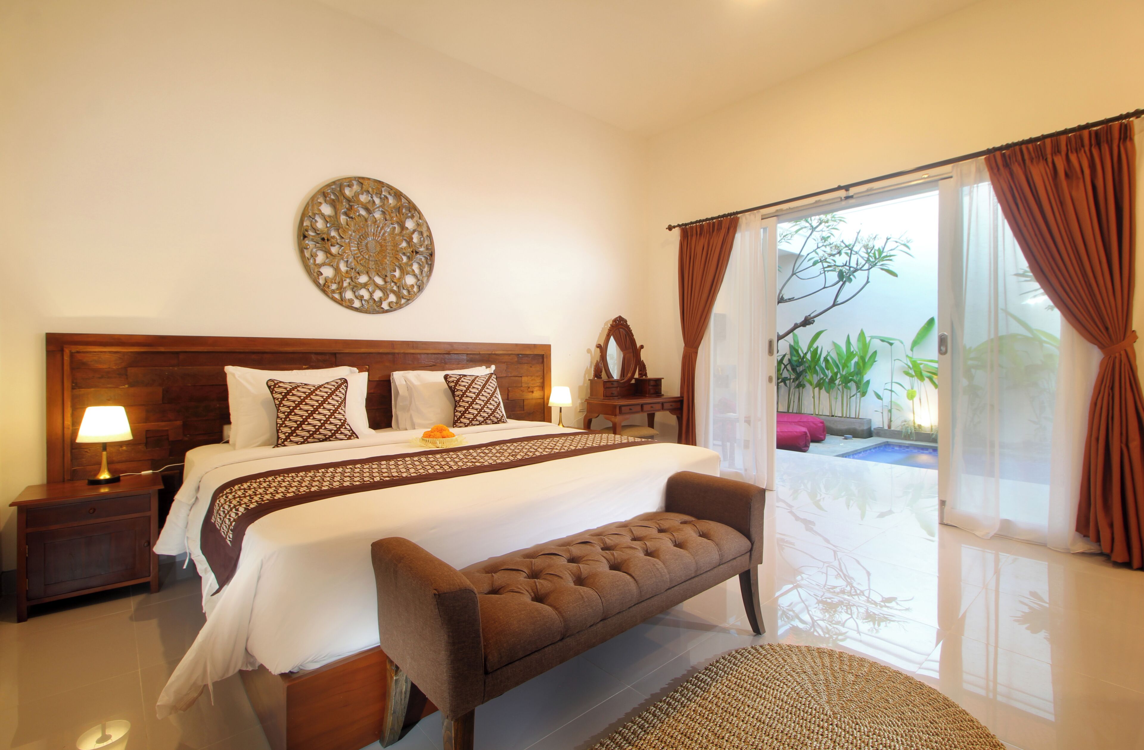 A bedroom with a large bed, a bench, a nightstand, a lamp, a decorative wall piece, and a view of a pool through a sliding door.