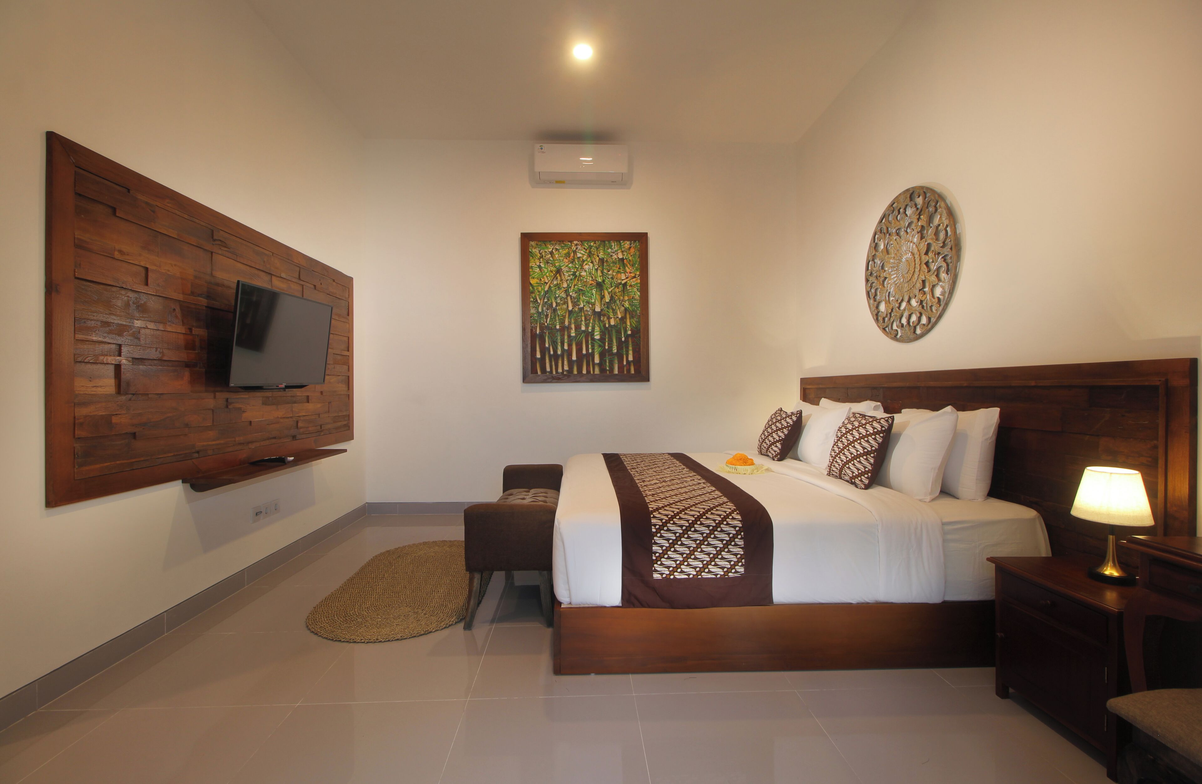 A bedroom with a wooden headboard, a flat-screen TV mounted on the wall, a bed with patterned bedding, a bedside table with a lamp, and a decorative wall piece.
