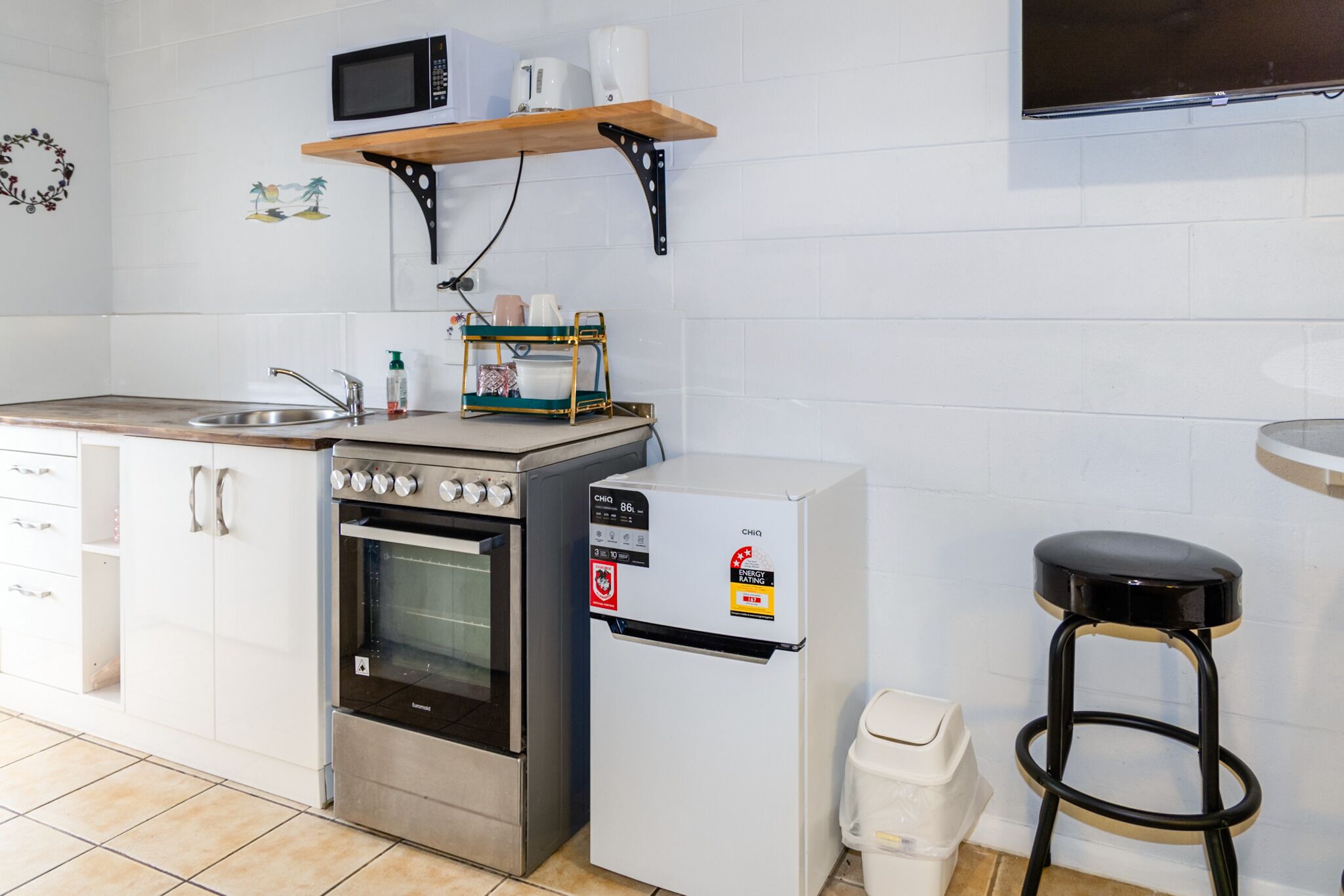 A compact kitchen with a built-in oven, a refrigerator, a microwave, and a sink. There is a wooden shelf above the microwave and a small stool to the right.