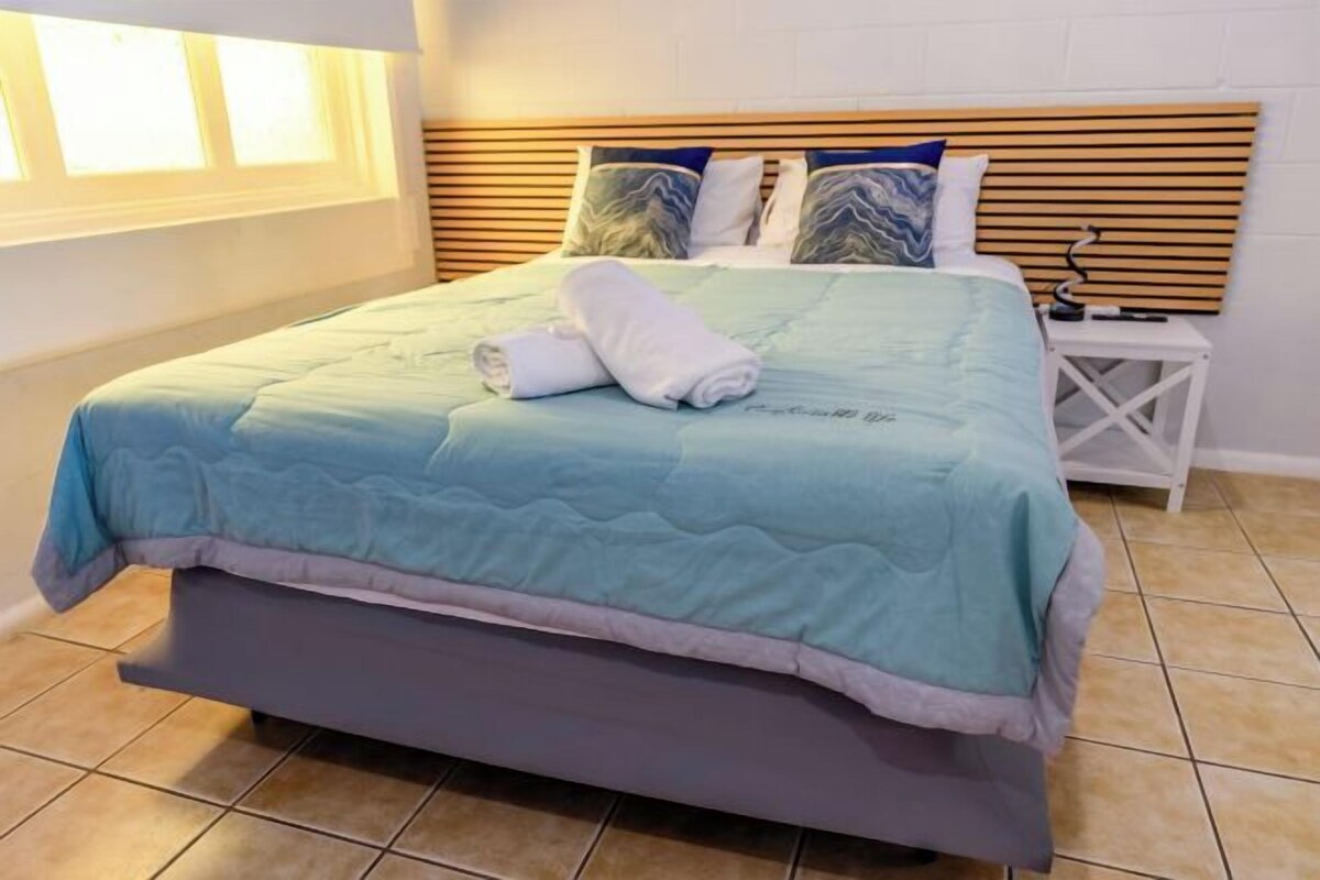 A neatly made bed with a blue and white quilt, a wooden headboard, and a white nightstand with a lamp.
