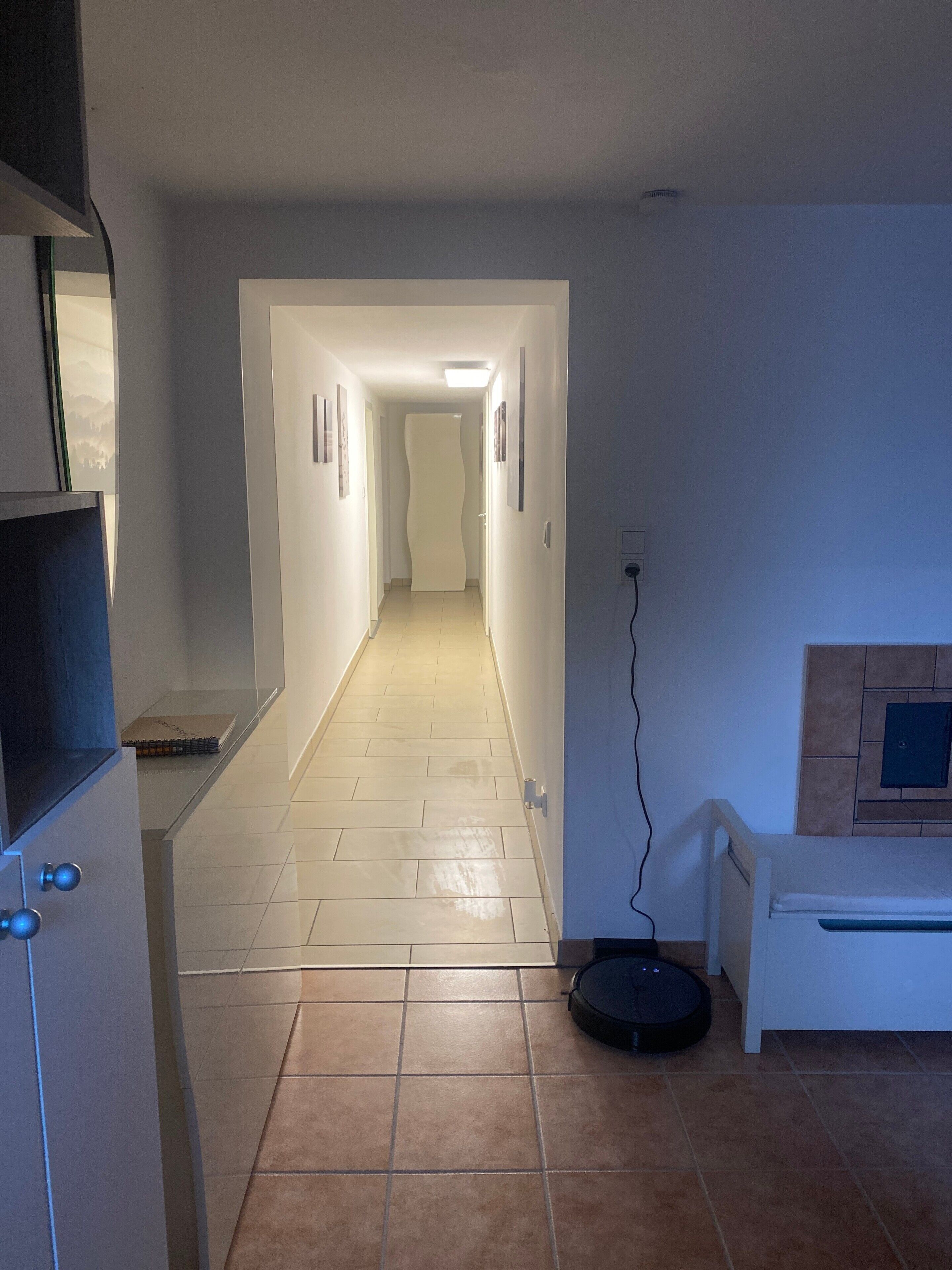 A narrow hallway with a tiled floor, a door at the end, and a wall-mounted electrical outlet.