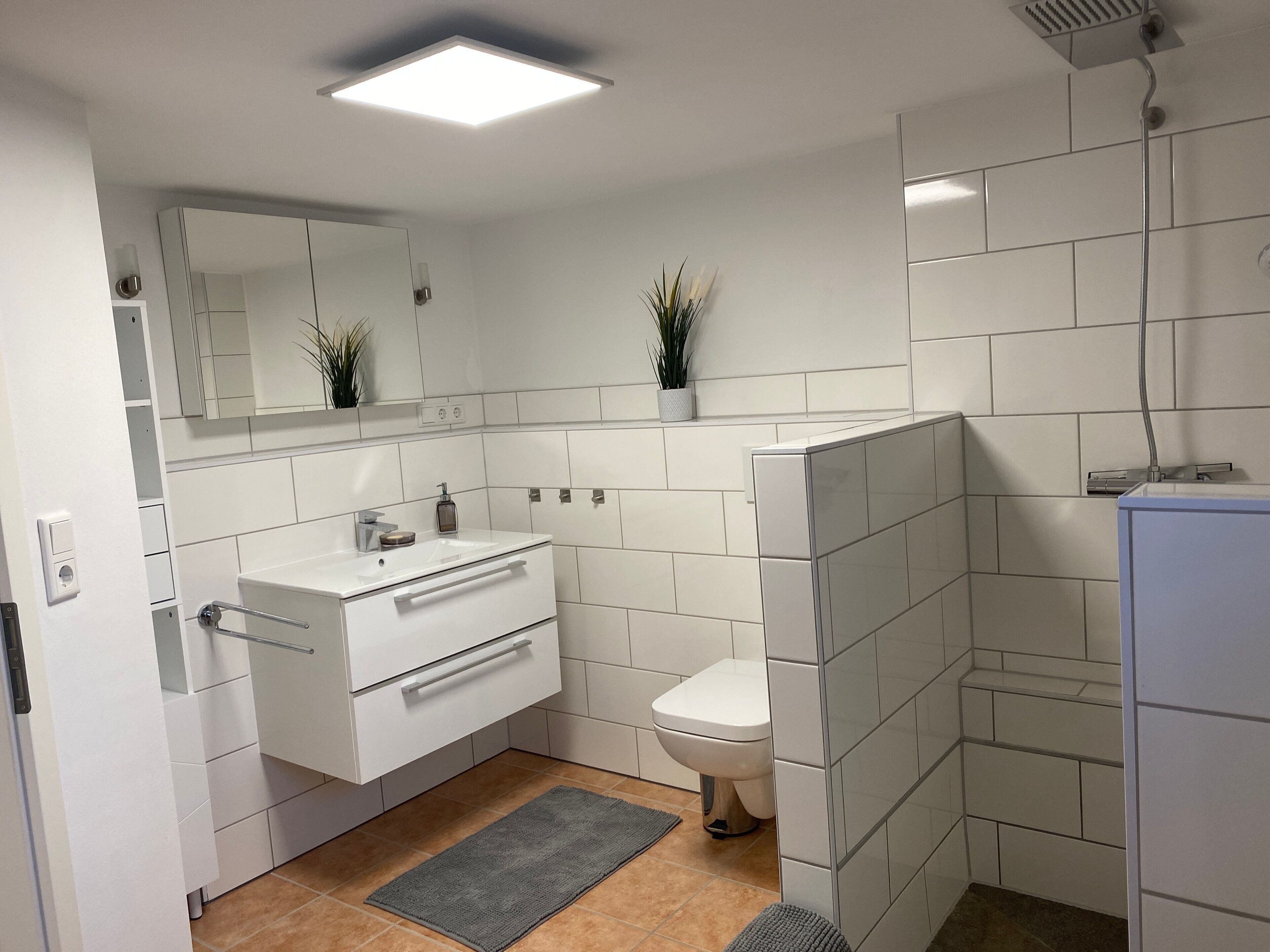 Un bany modern amb rajoles blanques, un lavabo blanc, un vàter, una dutxera i un mirall.