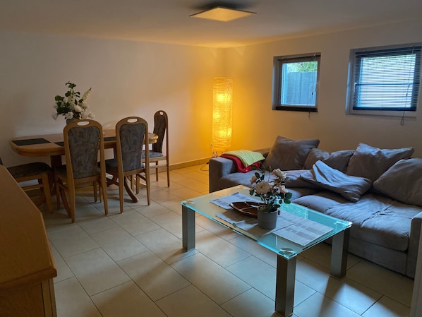 Ein Wohnzimmer mit Glastisch, einer Couch, einem Esstisch mit Stühlen und einem Fenster mit Jalousien.