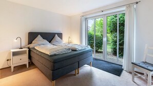 A bedroom with a bed, a nightstand, a chair, and a view of greenery through a sliding door. - Holiday Home 'Ferienhaus Sprante' with Private Terrace, Private Garden and Wi-Fi (Brunsbüttel)