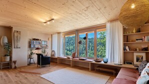 A cozy room with wooden floors, a large window with a view of trees, a bookshelf, and a comfortable seating area. - Apartment 'Am Stadtwald' with Mountain View, Private Terrace and Wi-Fi (Triberg)