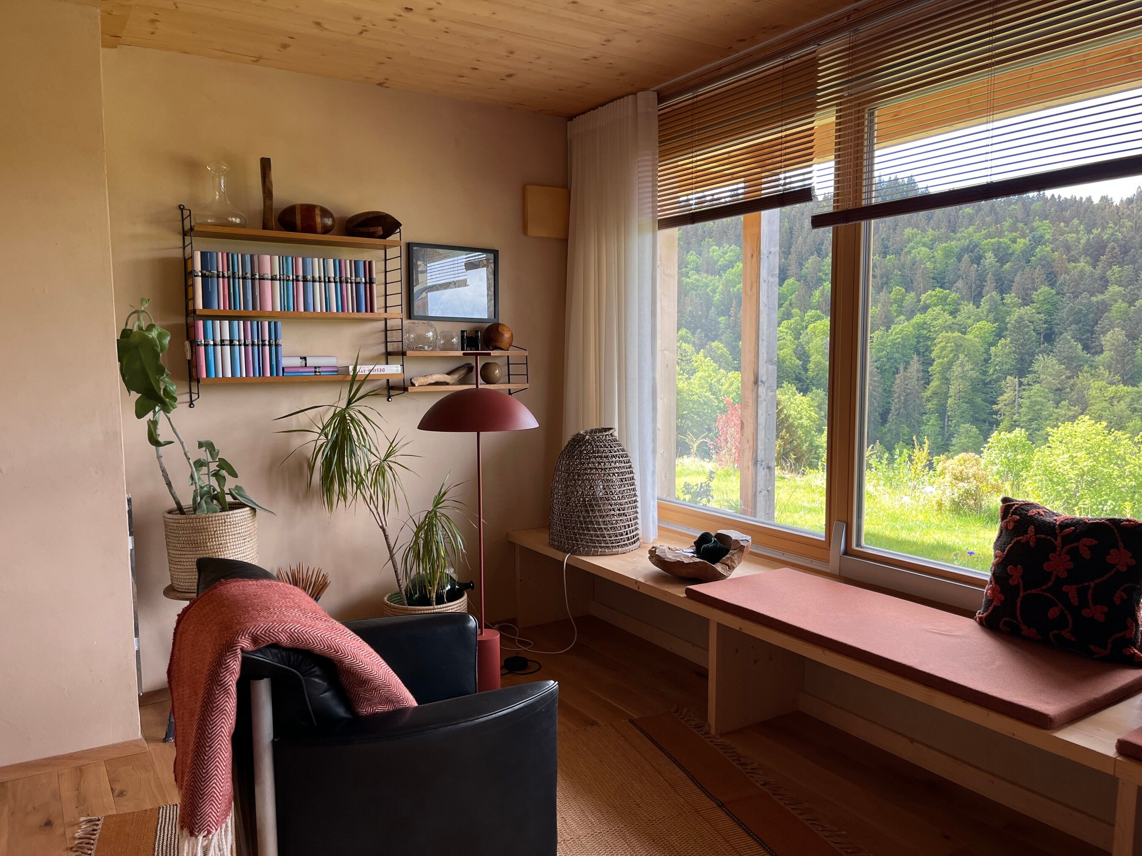 A cozy living room with a wooden ceiling, a bookshelf, a desk, a comfortable chair, a bench with a view of a forest, and a large window with blinds.