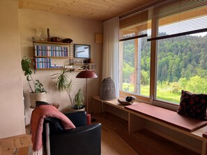 Een gezellige woonkamer met een houten plafond, een boekenkast, een bureau, een comfortabele fauteuil, een bank met uitzicht op een bos, en een groot raam met zonwering.