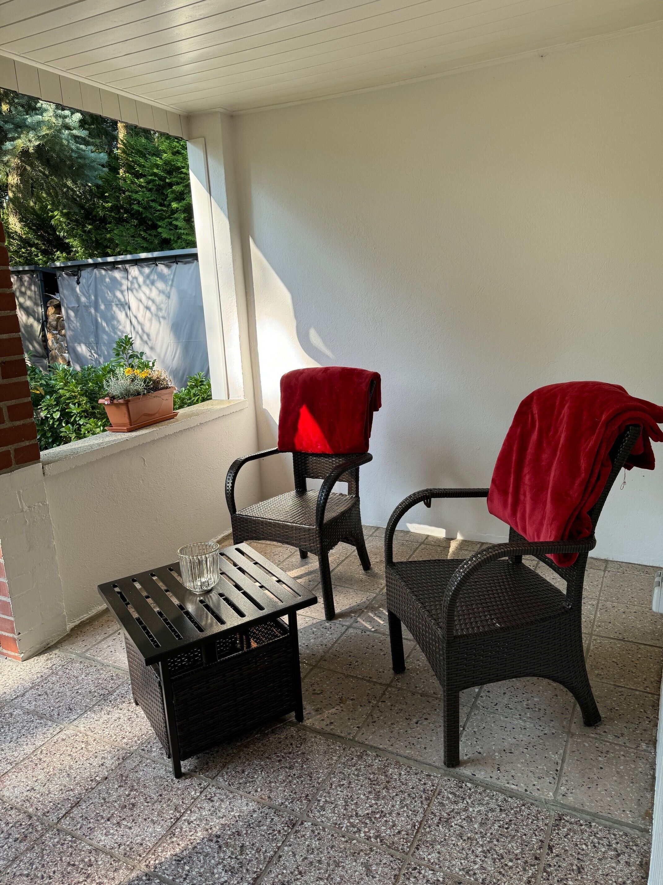 A covered patio area with two wicker chairs, one with a red cloth, a small table with a glass, and a potted plant.