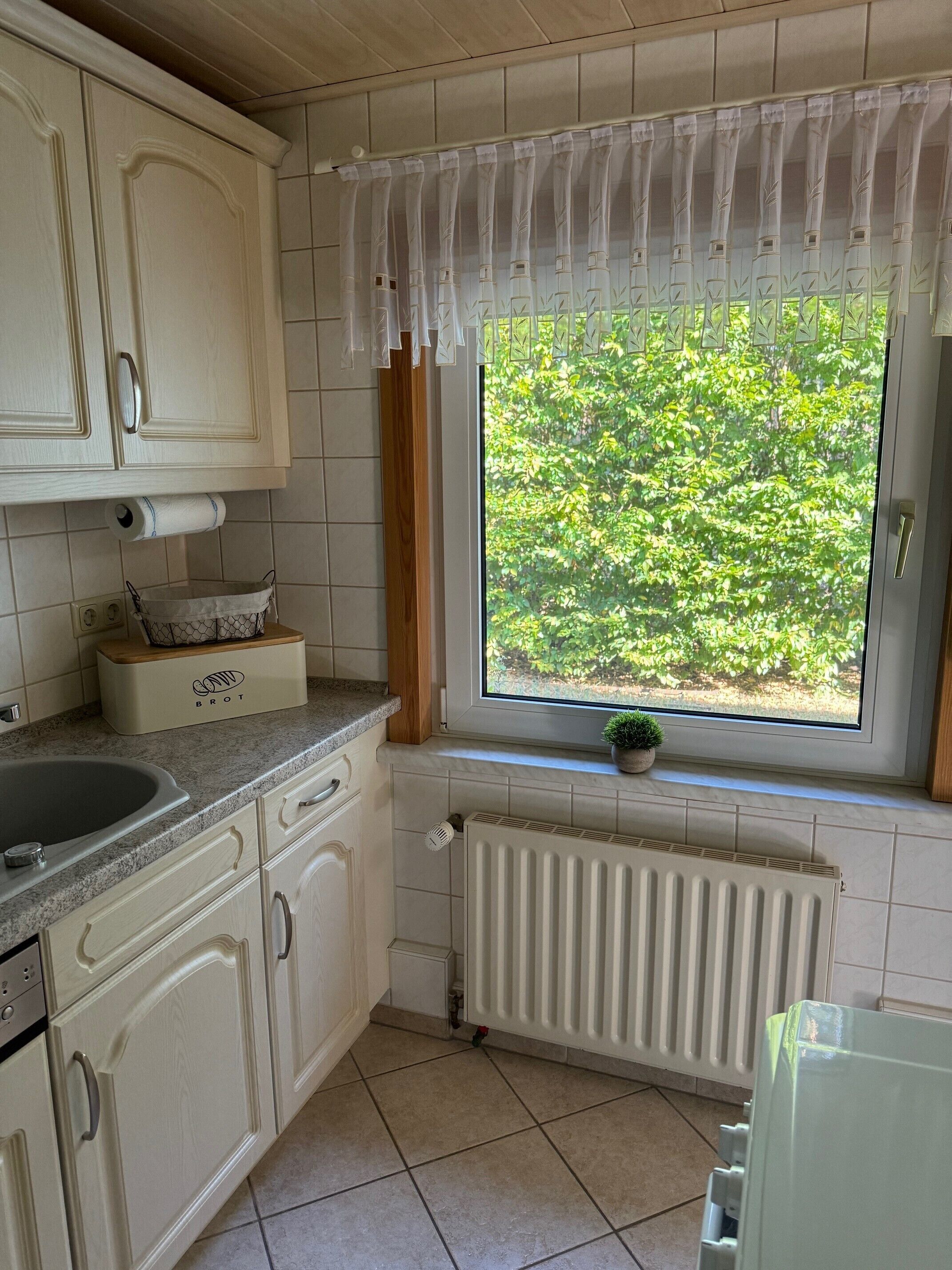 Una cocina con armarios blancos, un ventanal con vistas a zonas verdes, un radiador y una encimera con fregadero.
