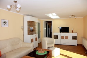 A living room with a sofa, a coffee table, a TV on a cabinet, a ceiling fan, and a door leading to another room.
