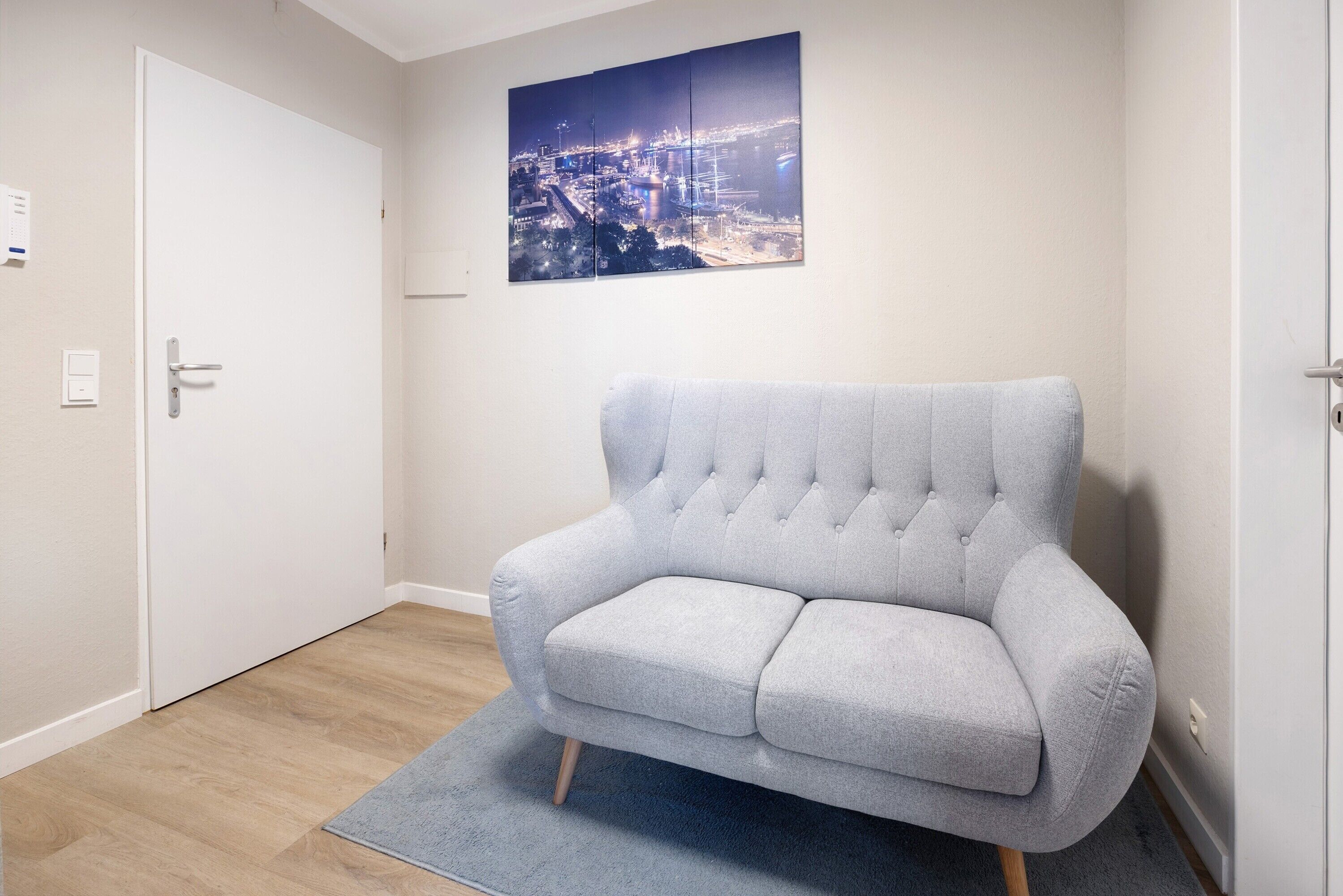 A room with a grey upholstered armchair, a light blue rug, a white door, and a cityscape painting on the wall.