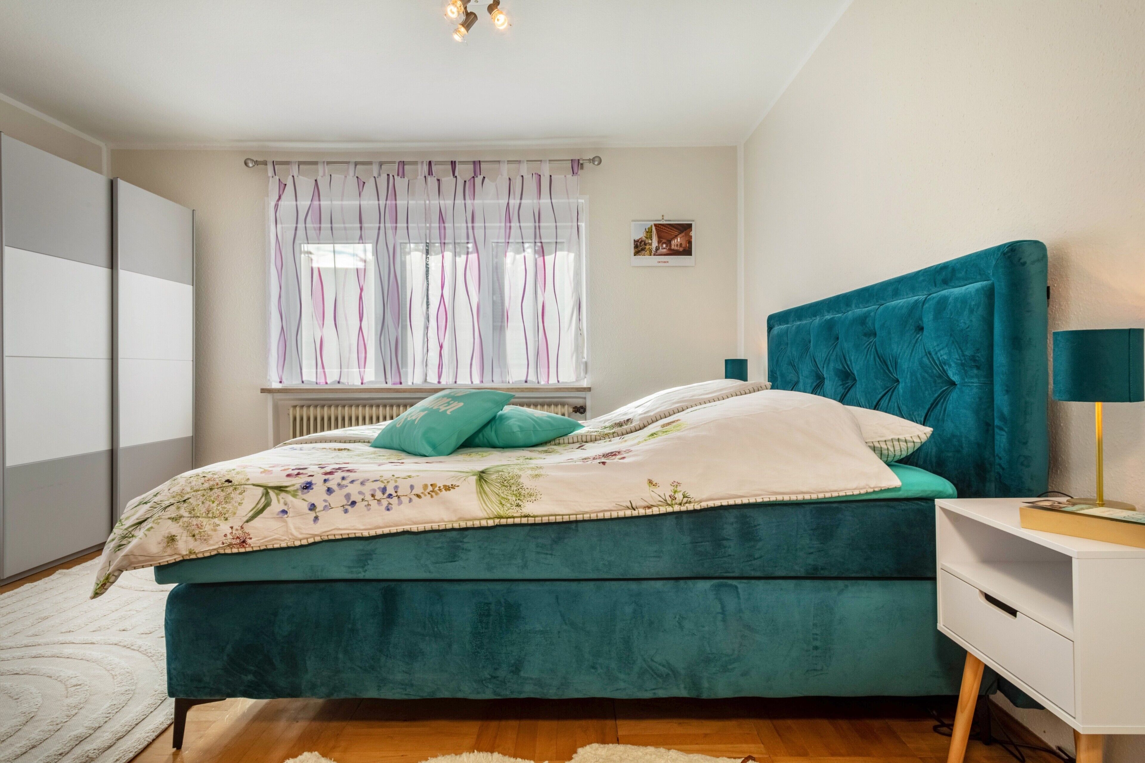 Une chambre  à coucher avec un lit de couleur turquoise, une table de chevet blanche et une fenêtre avec des rideaux.